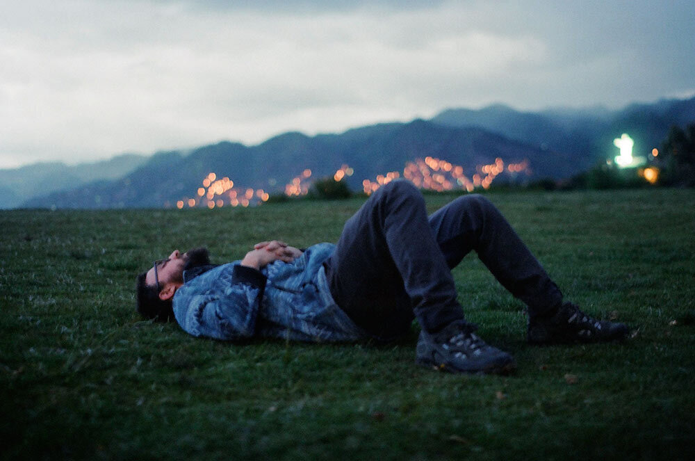 Matt laying on a grassy lawn looking up at a cloudy sky with Cusco, Peru in the background